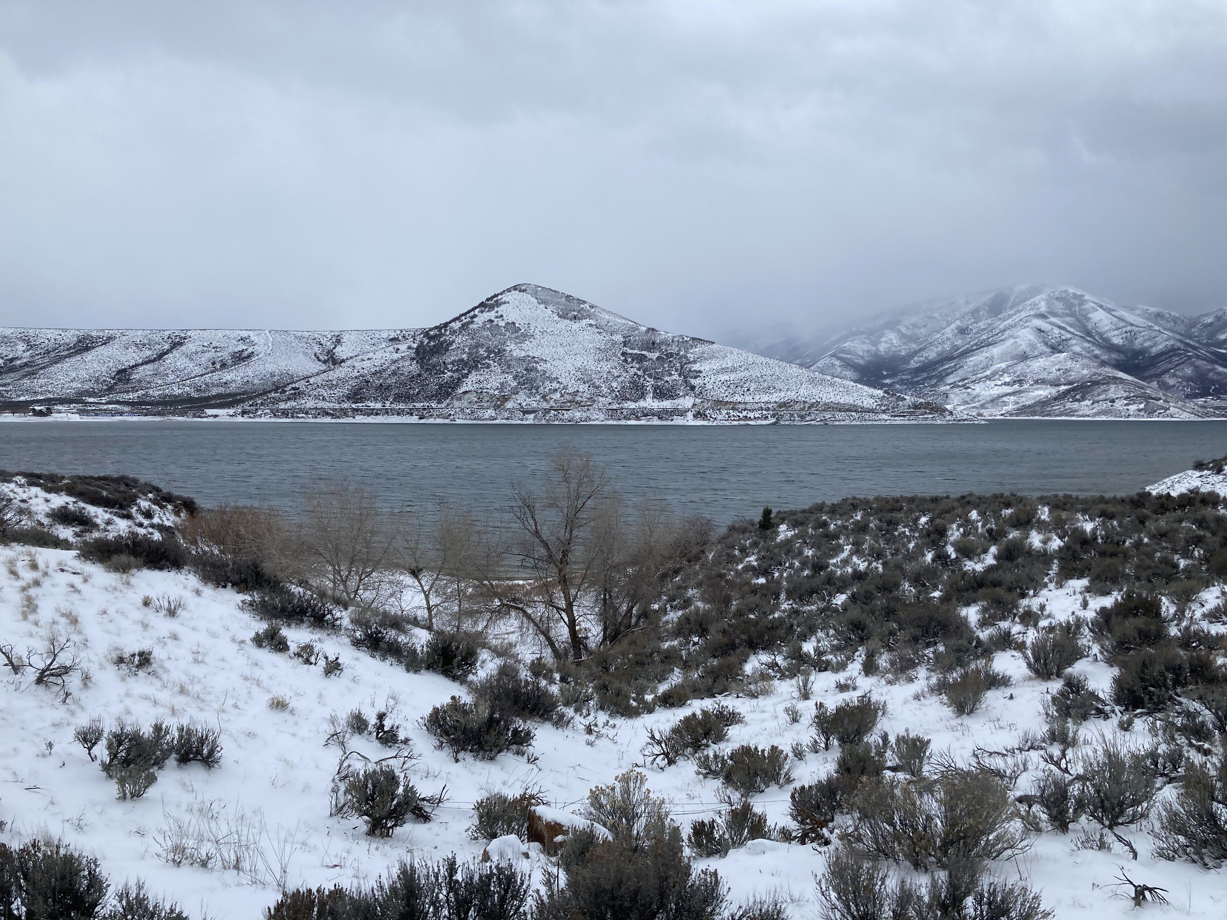 Deer Creek Reservoir under gray clouds, with snow on the sagebrush and the mountains behind