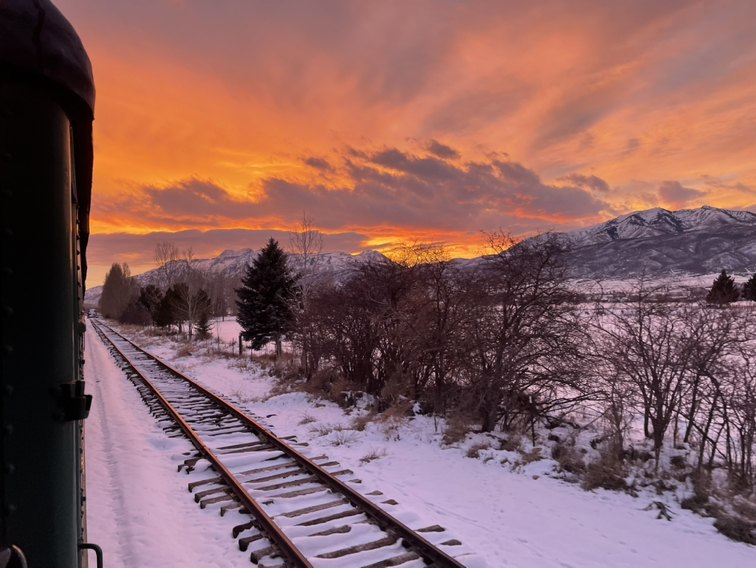 A winter sunset over snowy fields and the Wasatch, seen from the side of the train with the track running ahead