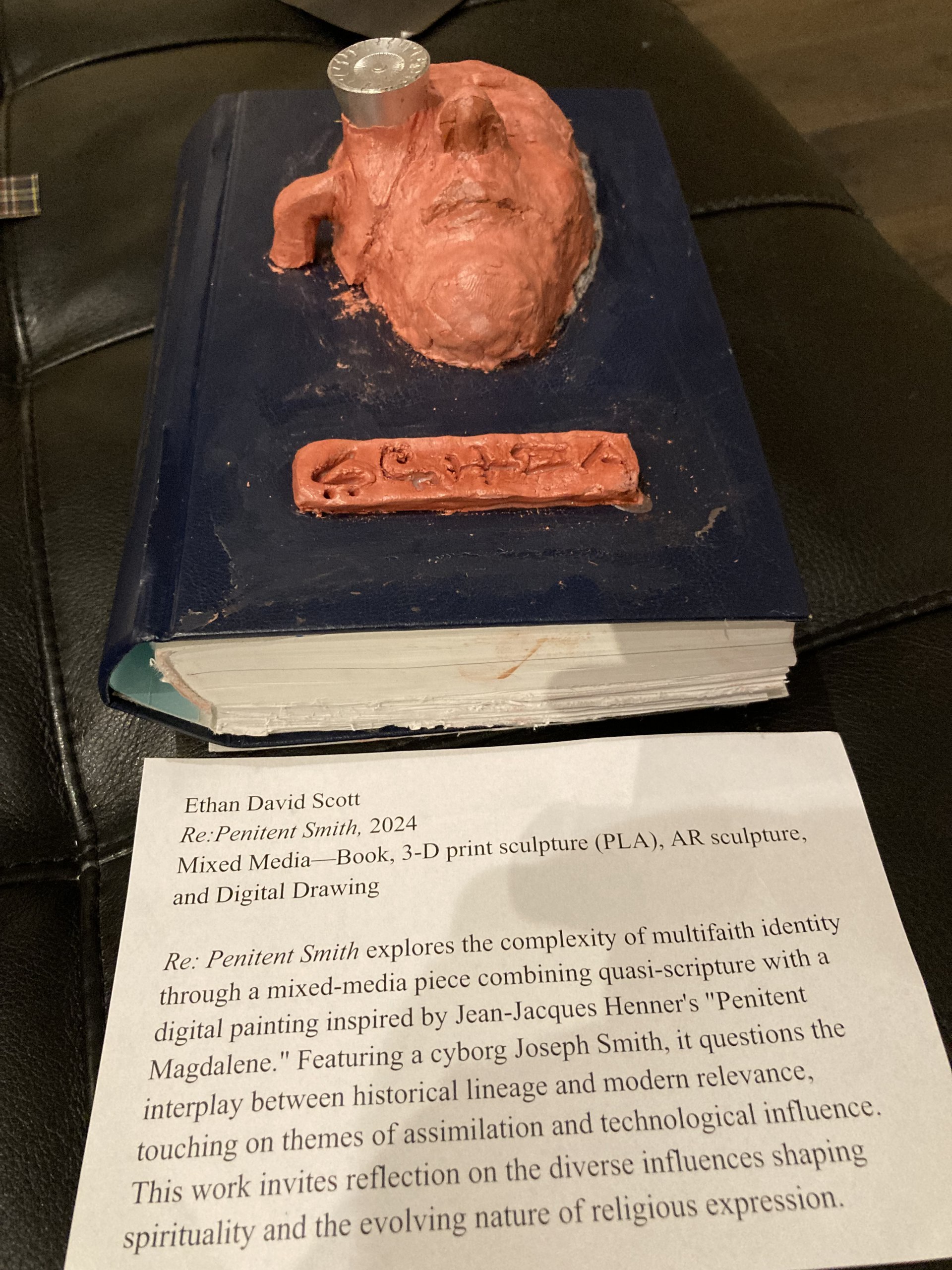 Re: Penitent Smith, a mixed-media sculpture: a clay face on a book, with its gallery card